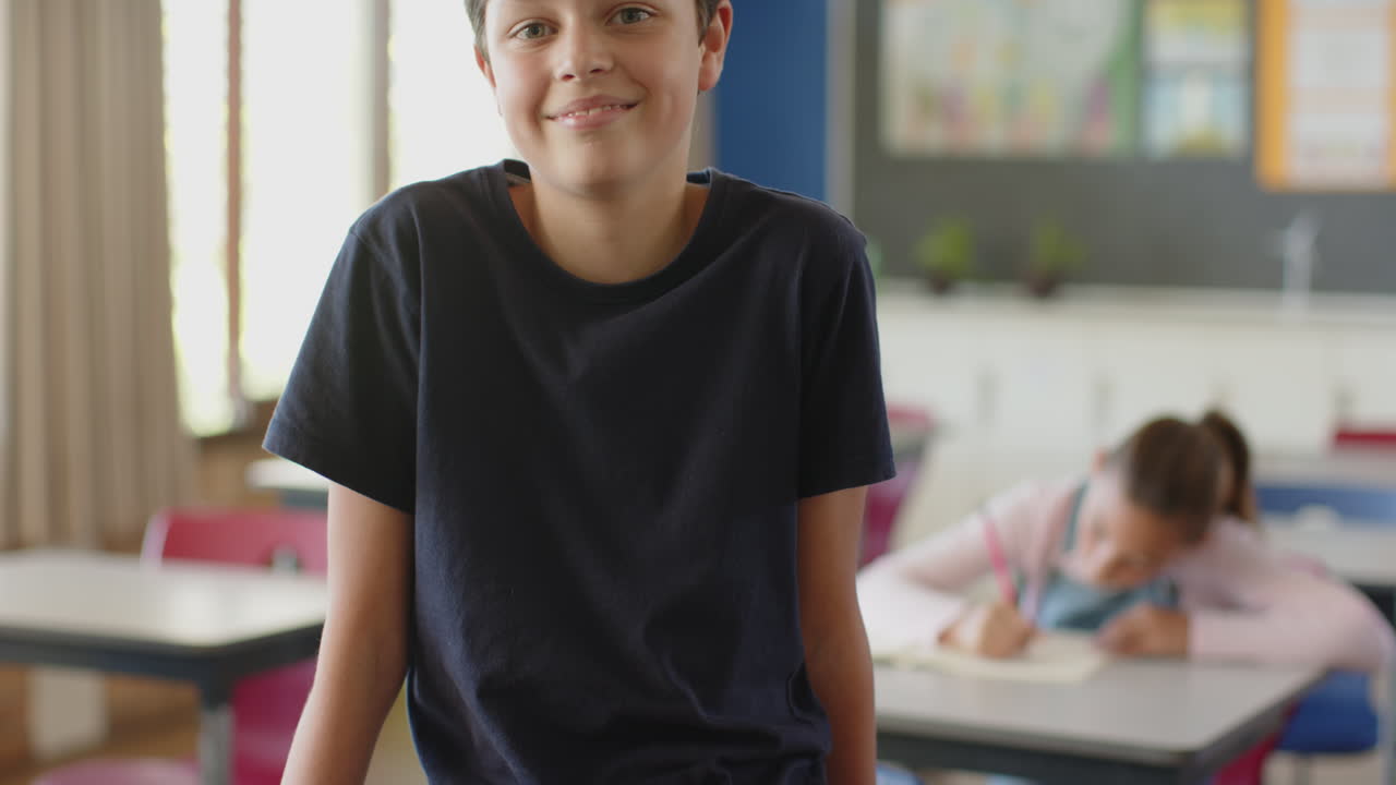 Ragazzo sorridente in un'aula scolastica, con una ragazza che scrive sullo sfondo, felice.