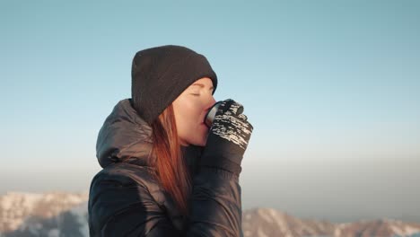 young caucasian woman with a winter jacket and beanie sipping tea out of a thermoflask in the mountains