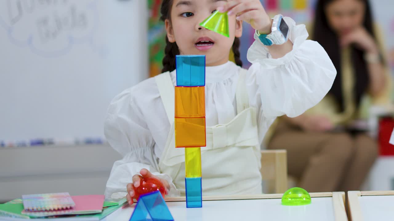 Young girl constructs tall tower with magnetic blocks in bright classroom, teacher observing, natural lighting