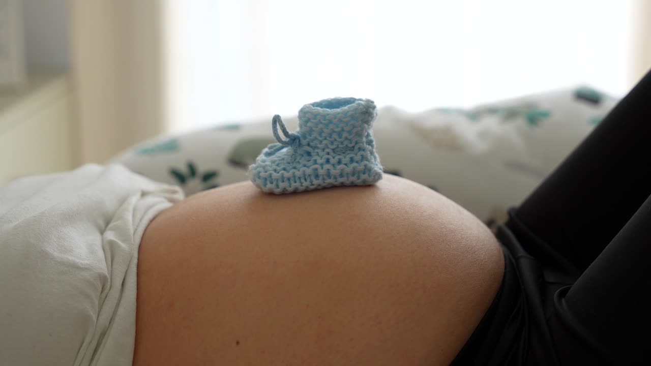 Blue knitted baby booties rest on pregnant belly as woman lies in bed, belly moves in breathing rhythm