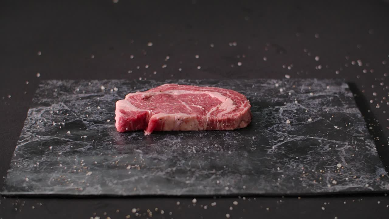 A fresh raw marbled ribeye beef steak falls onto a seasoned black marble board in slow motion against a dark studio background
