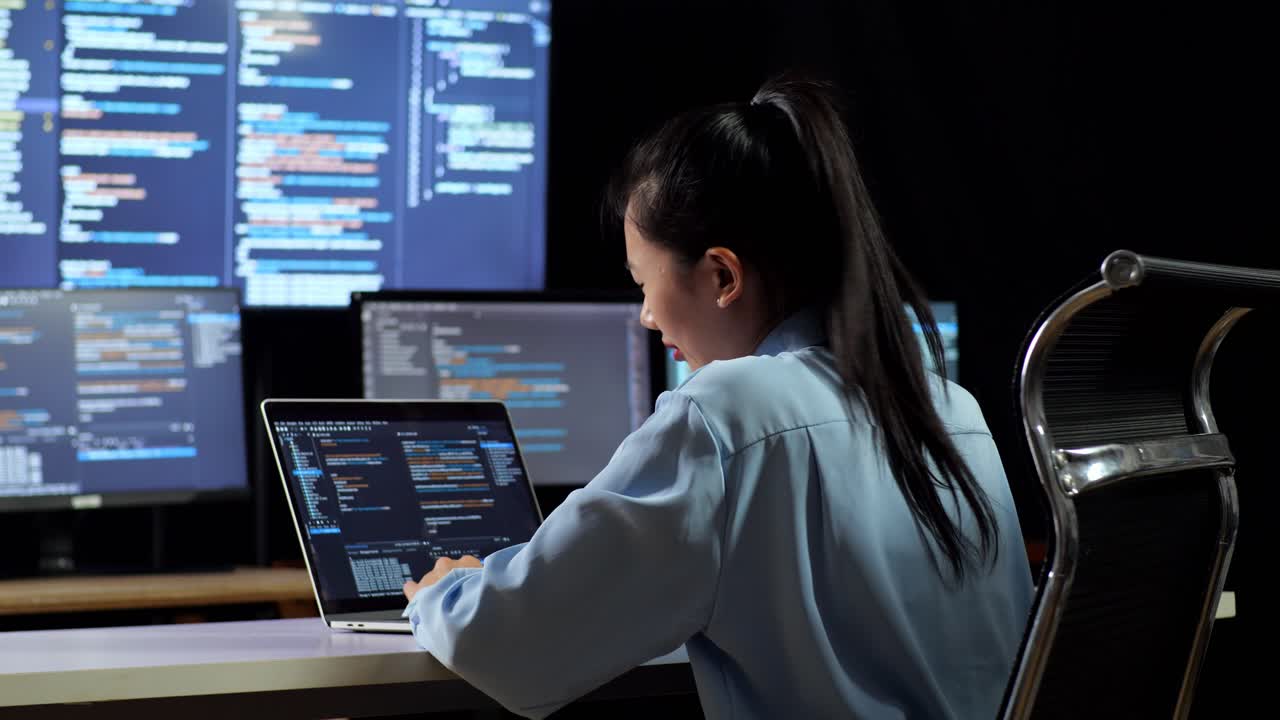vista trasera de una programadora asiática que tiene dolor de cabeza mientras escribe código en una computadora portátil usando múltiples monitores que muestran la base de datos en los escritorios de ventana de terminal en la oficina
