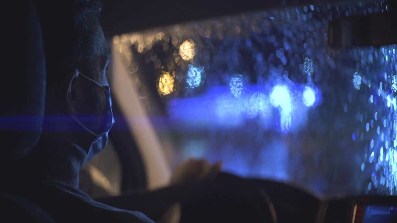 The young man in medical mask is sitting in the car on the rainy evening