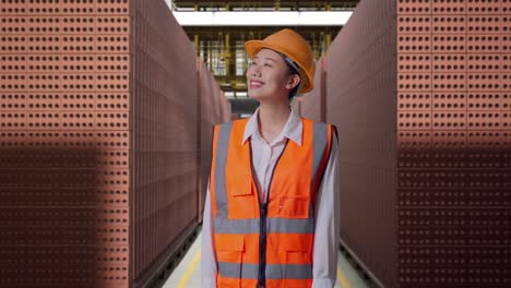 asian female engineer with safety helmet looking around while standing with red brick packed in stacks are stored