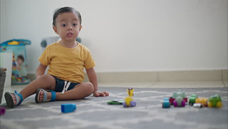 Young-cute-latin-toddler-sitting-on-the-floor-on-a-grey-carpet-making-faces-and-talking-to-the-camera