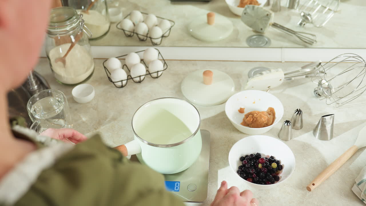 Rear view of lady placing pastel green pot with wooden handle on digital kitchen scale while turning device on, surrounded by baking ingredients like eggs, flour, berries, and kitchen utensils