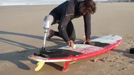 long shot of a male surfer with prosthetic leg waxing surface of surfboard