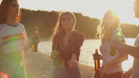 tres chicas calientes están bailando con camisetas cortas y pantalones cortos con cerveza en la playa de arena al atardecer. su cabello está volando en el viento.