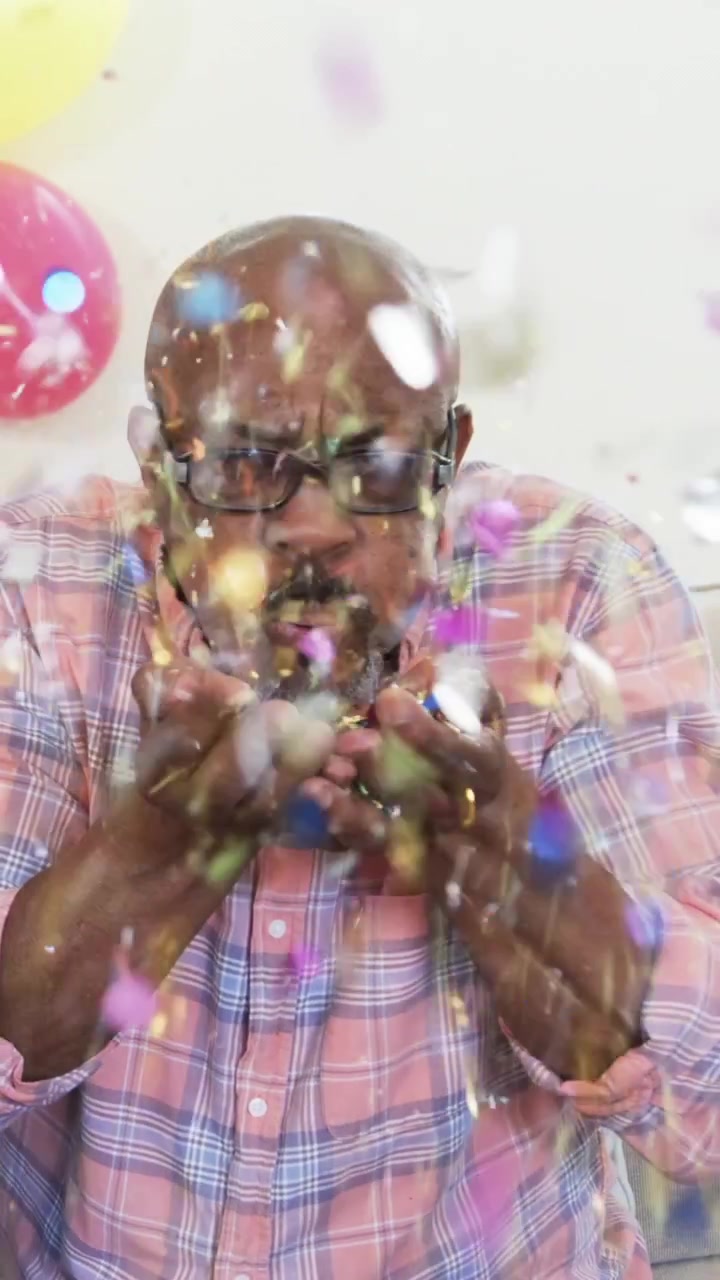 Video verticale: Uomo afroamericano anziano che soffia coriandoli dalle mani per divertimento sul divano con palloncini.