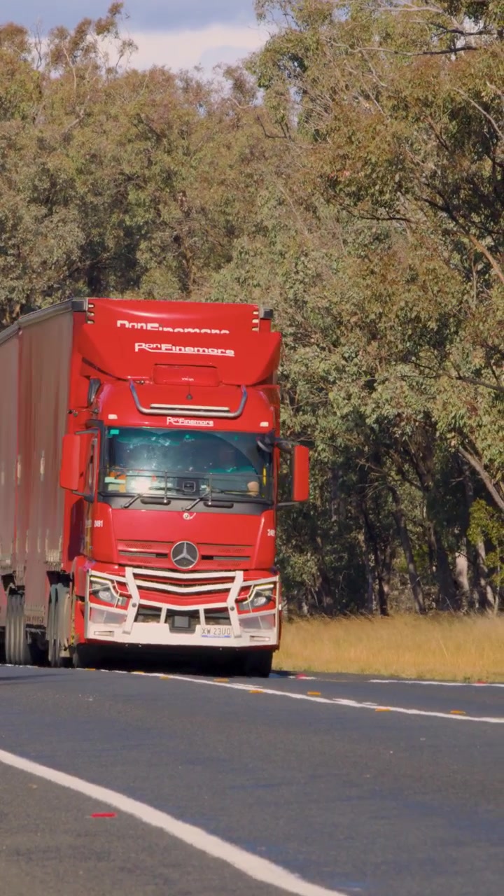 Due grandi autotreni viaggiano lungo una strada rurale assolata, costeggiata da alberi. Una telecamera fissa riprende entrambi i camion in sequenza, in pieno giorno, con luce chiara e brillante.