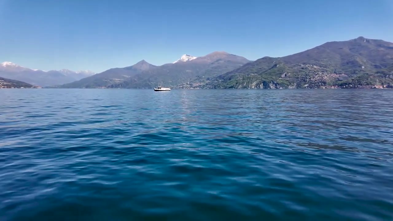 Gita panoramica in traghetto attraverso il Lago di Como in Italia, da Varenna a Menaggio, con acqua blu vivido, barche in lontananza, cime alpine innevate e paesaggi montani panoramici.