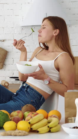 happy girl eats salad in a white kitchen near a table with fruits and vegetables. vertical video
