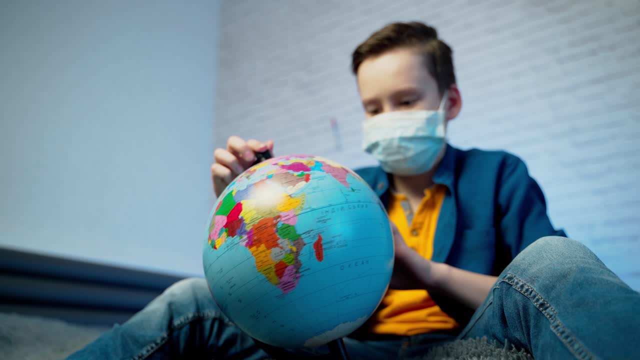 Boy looking at globe learning about virus spreading around the world. Boy wearing medical face mask. Concept of COVID-19 pandemic infection.