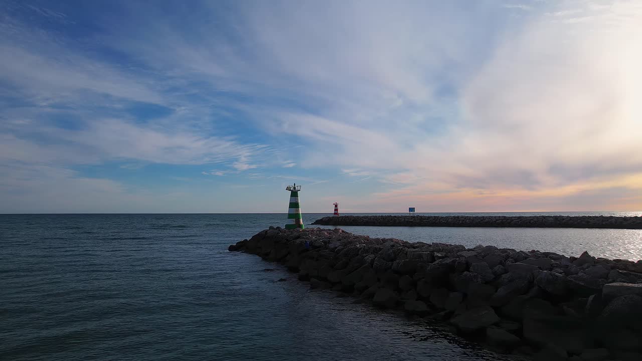Drone tracking shot along the green harbour beacon on the outer breakwater at the entrance to Marina de Vilamoura, with stripped lighthouse and Atlantic Ocean beside Vilamoura Beach in Quarteira