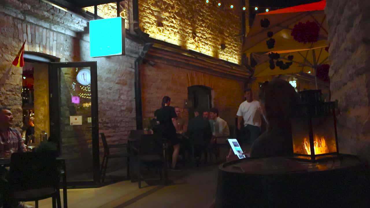 Narrow street filled with people at Rotermanni block in Tallinn during night or evening time while people eat, drink and chat. Limestone architecture that is well lit by lights, fire lantern in front