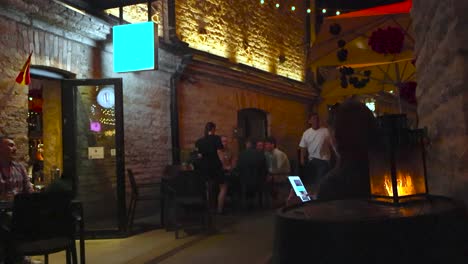 narrow street filled with people at rotermanni block in tallinn during night or evening time while people eat, drink and chat. limestone architecture that is well lit by lights, fire lantern in front