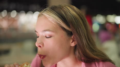 woman in pink dress takes a bite from burger while holding potato chips, eating with a satisfied expression, the background features a blurred view of a person in red walking by and blur light view