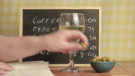 Hand-reading-book-with-glass-of-white-wine-and-green-olives-in-cafe-medium-shot