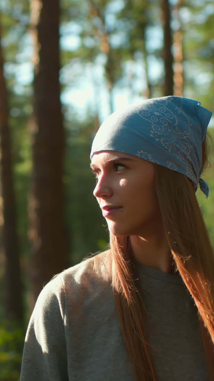 две подруги прогуливаются по солнечному лесу, одна в синем платке, а другая в красном рюкзаке, теплые улыбки и нежные выражения лица, когда солнечный свет проходит через высокие деревья.