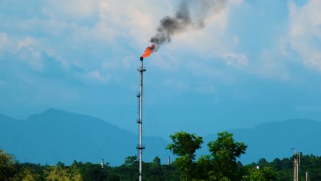 industrial flare stack burning gas with smoke against a mountain backdrop