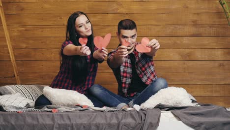 beautiful couple are sitting on the bed and showing red hearts by handmade in their hands on valentine's day