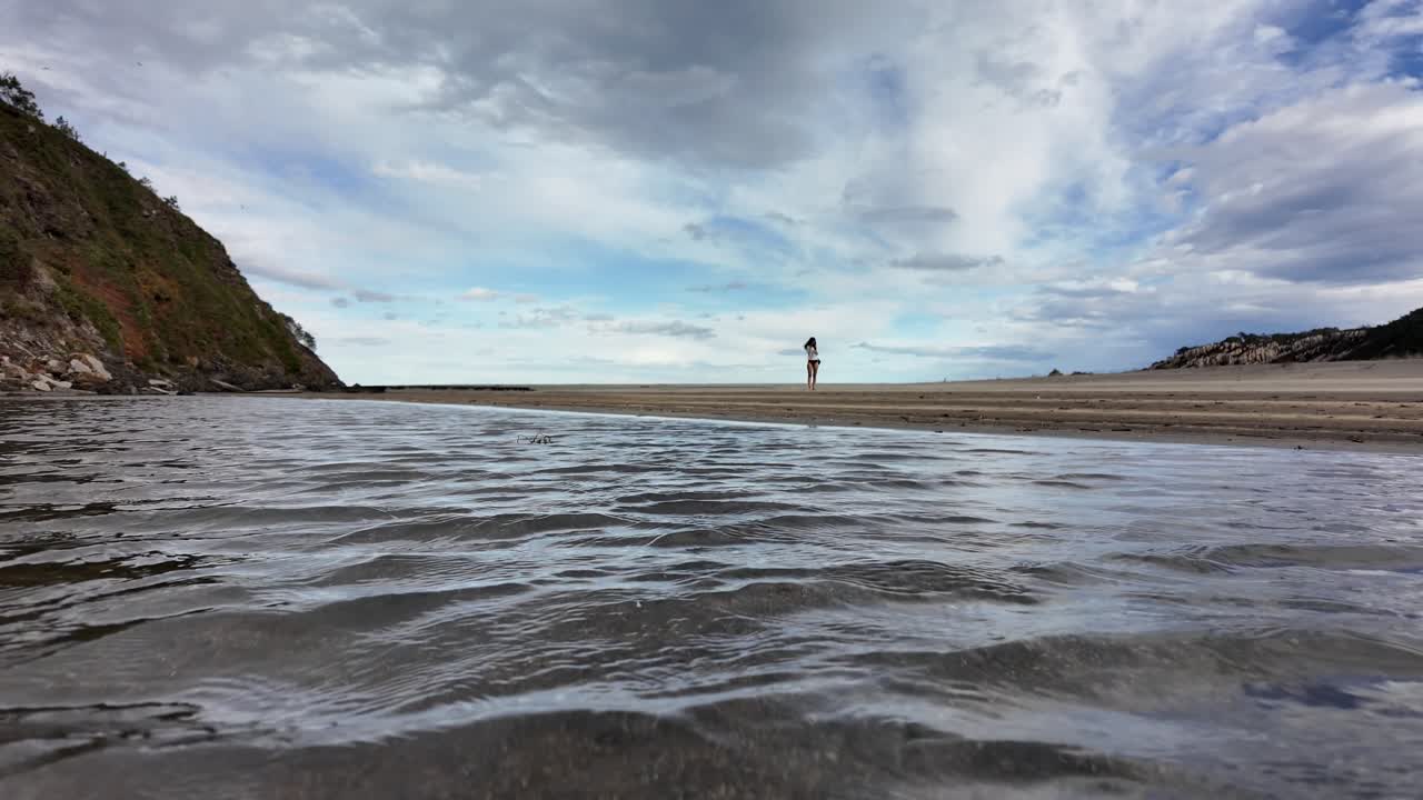 A low-angle shot from the water shows gentle ripples and a distant person standing on a wide sandy beach