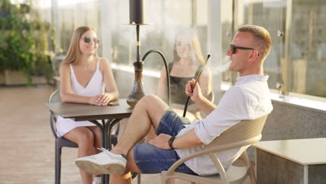 relaxed man sitting in the company of two girls smoking hookah. young people are chatting enjoying time in a café.