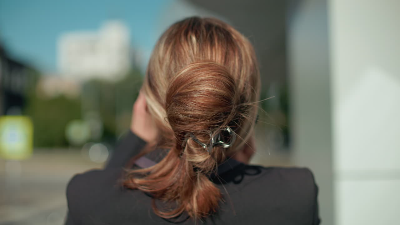 Close up rear view of woman in black suit with hair tied back sipping coffee on phone call as she walks in urban setting near glass building with blur background and soft natural light