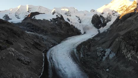 Sobrevuelo-Aéreo-Sobre-El-Final-Del-Glaciar-Morteratsch-En-Engadin,-Suiza-Al-Amanecer-Con-Una-Panorámica-Desde-Algunos-De-Los-Picos-Más-Altos-De-Los-Alpes-Suizos-Como-Piz-Bernina,-Piz-Palu