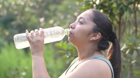 Lateinische-Frau-Trinkt-Wasser-Aus-Der-Flasche-Beim-Wandern-Auf-Dem-Von-Bäumen-Gesäumten-Fußweg-Des-Parks