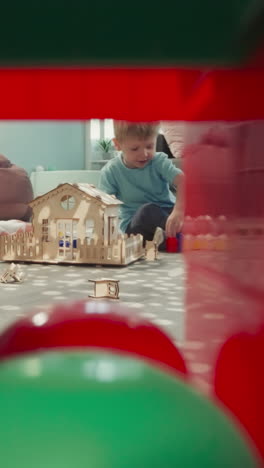 little boy builds wall from colorful blocks sitting near handmade wooden house. toddler plays with toys view through clearance in construction of cubes