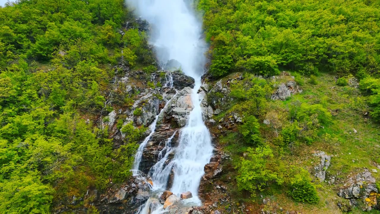 Vapore acqueo che si innalza da una cascata di montagna su terreno roccioso. Vista ravvicinata di spruzzi bianchi che si riversano su pietre grigie, circondati da un fogliame verde brillante.