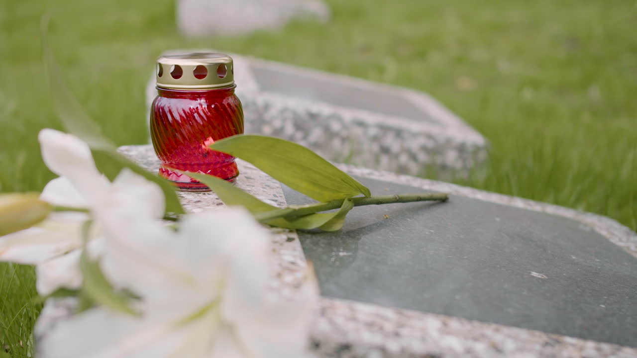 Close Up View Of Unrecognizable Man In Black Suit Kneeling And Putting Flowers And A Candle On Tombstone In A Graveyard