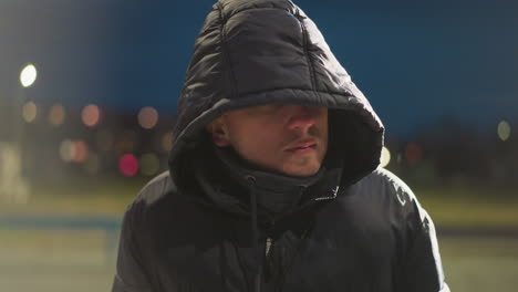 un hombre se sienta al aire libre por la noche en un estadio, apareciendo pensativo, con una capucha cubriendo su cabeza, el fondo presenta una vista borrosa de edificios y luces oscuras