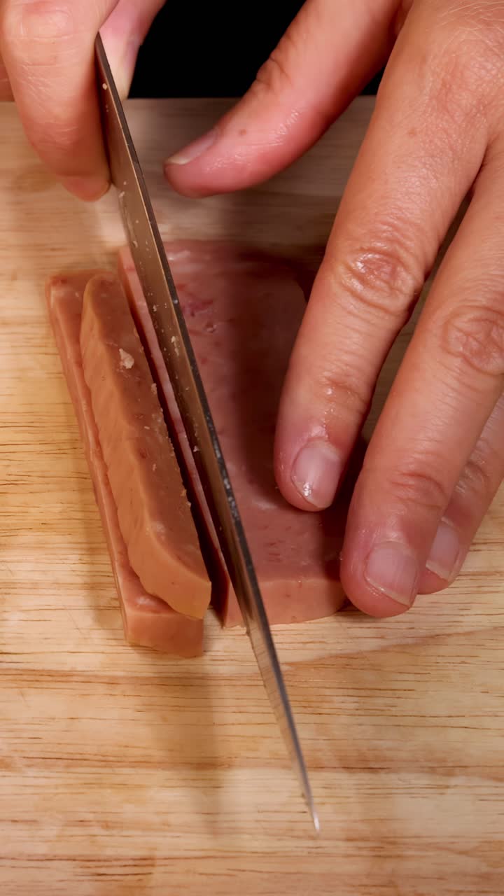 Una mano utiliza un cuchillo de cocina afilado para cortar fiambre procesado en tiras uniformes sobre una tabla de cortar de madera, bajo una iluminación brillante y directa, en un primer plano cenital.