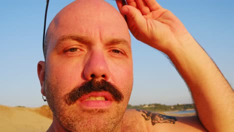 hombre caucásico con alopecia calva con bigote usando gafas de sol en la playa durante la puesta de sol