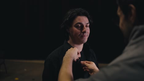 Over-the-shoulder-a-confident-male-stage-director-in-gray-clothes-attaches-a-small-microphone-to-a-young-brunette-actor-in-a-black-suit-before-going-on-stage-in-a-theater-during-a-performance