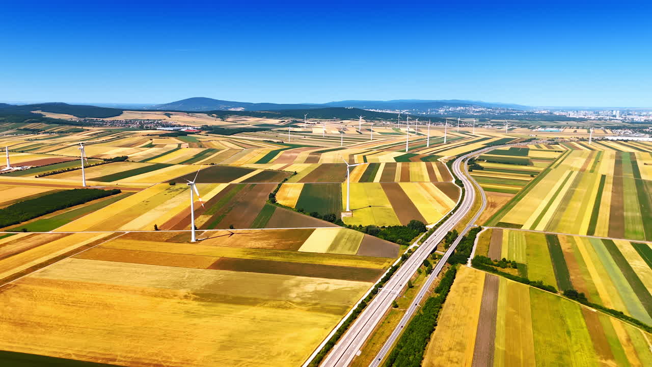 Beautiful vast agricultural fields of colorful patches crossed by the highway. Wind turbines are scattered by the plantation. Aerial view