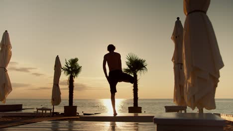 The-guy-stands-on-one-leg,-raises-his-hands-up,-and-then-lowers-them-on-the-beach-in-the-morning.-Sunrise-Meditation