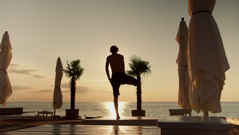 the guy stands on one leg, raises his hands up, and then lowers them on the beach in the morning. sunrise meditation