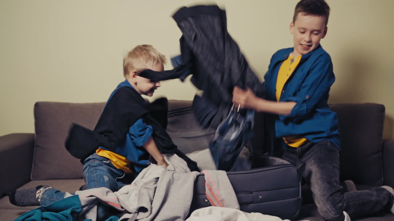 Smiling kids packing a grey suitcase with different clothes at home. Two cheerful brothers wearing blue shirts put things into the big valise in the room. Slow motion.
