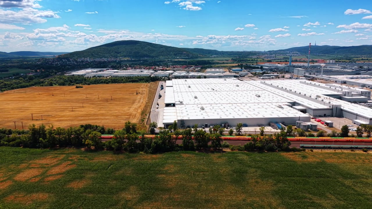 Capannone industriale accanto a un campo raccolto e una ferrovia. Un binario alberato con vagoni colorati separa le colture verdi dai tetti bianchi e dalla stoppia dorata