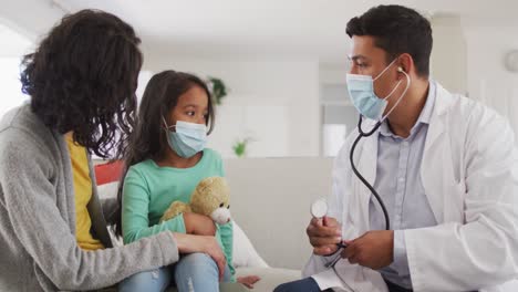 médico masculino hispano con un estetoscopio hablando con la madre y la hija en casa con máscaras faciales