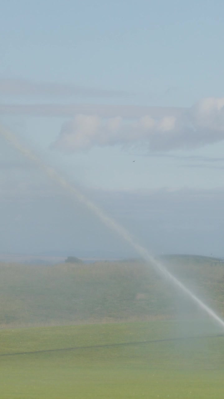 Un grande irrigatore irriga il green di un campo da golf in una giornata limpida. La telecamera rimane ferma, catturando lo spruzzo d'acqua, il paesaggio erboso e le colline distanti.