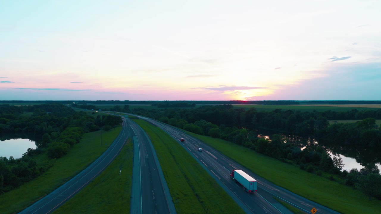 Ampia vista aerea con drone di un camion logistico bianco che trasporta merci lungo un'autostrada a più corsie attraverso un paesaggio verde lussureggiante con fiumi durante un tramonto dorato.