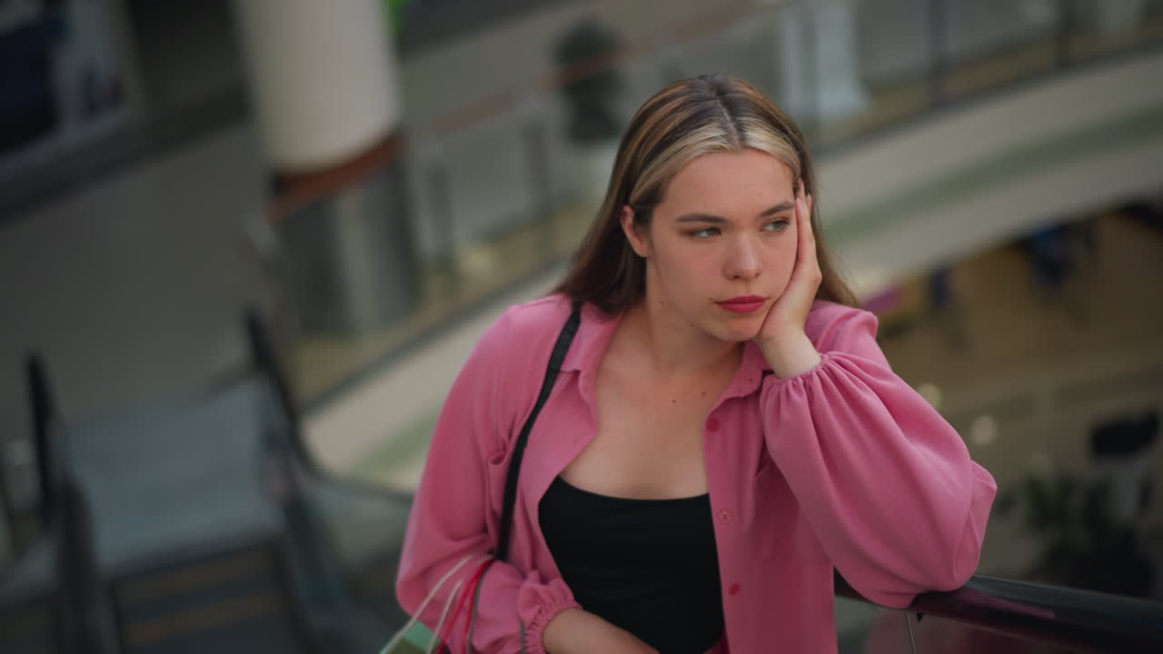 분홍색 옷을 입은 여자가 그녀의 에 손을 놓고, 사려 깊게 보이며, 오른손에 쇼핑 가방을 들고, 쇼핑몰의 흐릿한 배경으로 움직이는 에스컬레이터를 타고 있습니다.