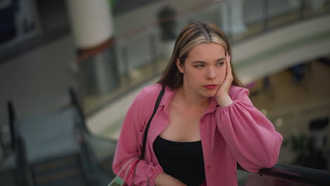 dama con tela rosa, coloca su mano en su barbilla, apareciendo pensativa, sosteniendo una bolsa de compras en su mano derecha, mientras monta en una escalera mecánica en movimiento con un fondo borroso de un centro comercial