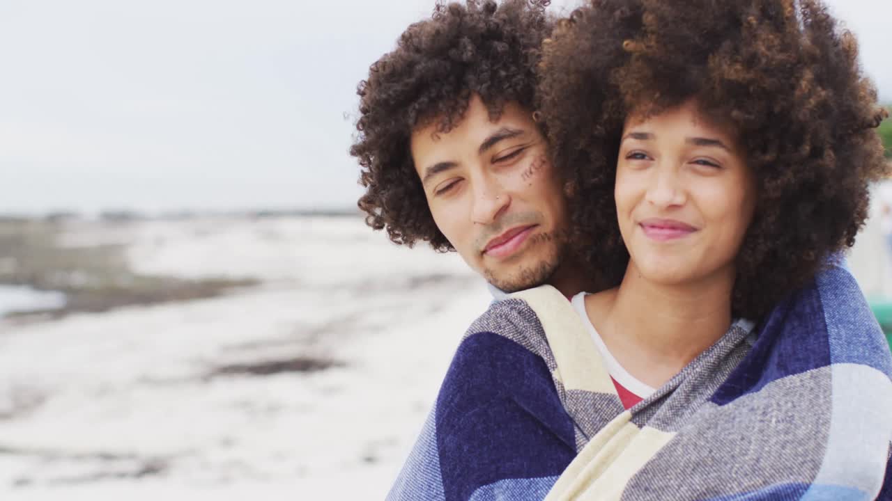 Close-up de casal afro-americano enrolado em cobertor, sorrindo no calçadão perto da praia.