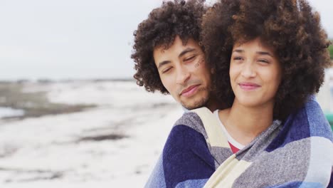 close up of african american couple wrapped in blanket smiling on the promenade near the beach