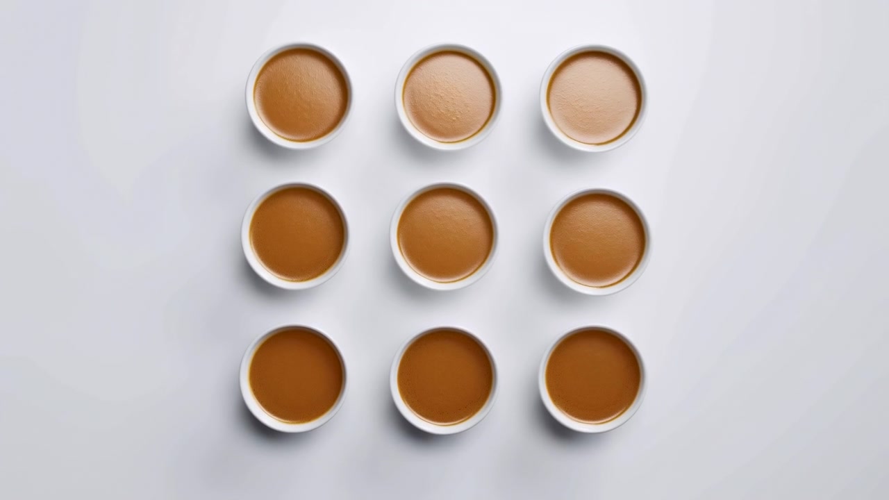 Nove tazze di ceramica bianca riempite fino all'orlo con un liquido liscio color caramello, disposte in una rigorosa griglia 3x3 su un tavolo grigio freddo, creando una composizione da studio minimale ed equilibrata con luce soffusa